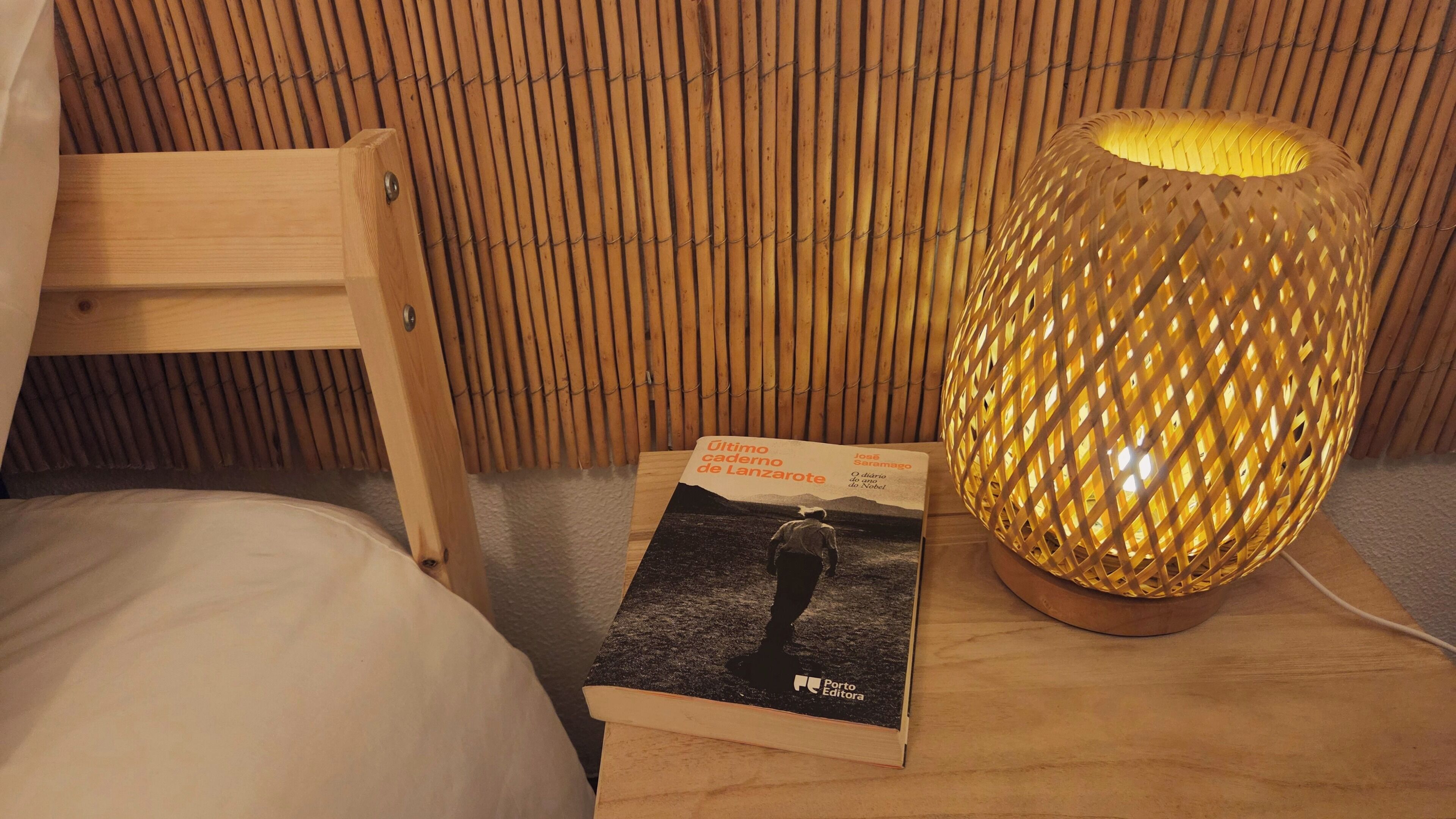 En seng med trægavestykke, et natbord med en vævet lampe, og en bog med titlen 'Último caderno de Lanzarote'.