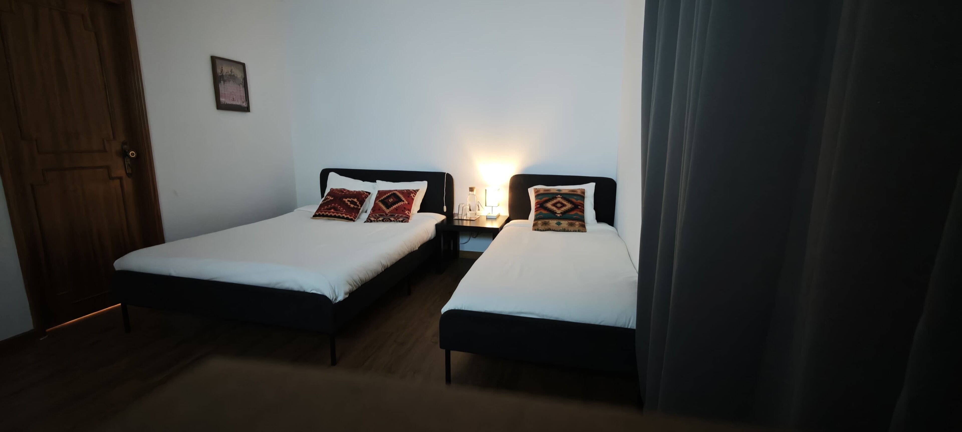 A hotel room with two single beds, a wooden door, a nightstand with a lamp, and a framed picture on the wall.