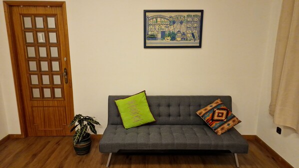 Ein Zimmer mit grauem Sofa, einer Holztür, einem gerahmten Bild an der Wand und einem Blumentopf.