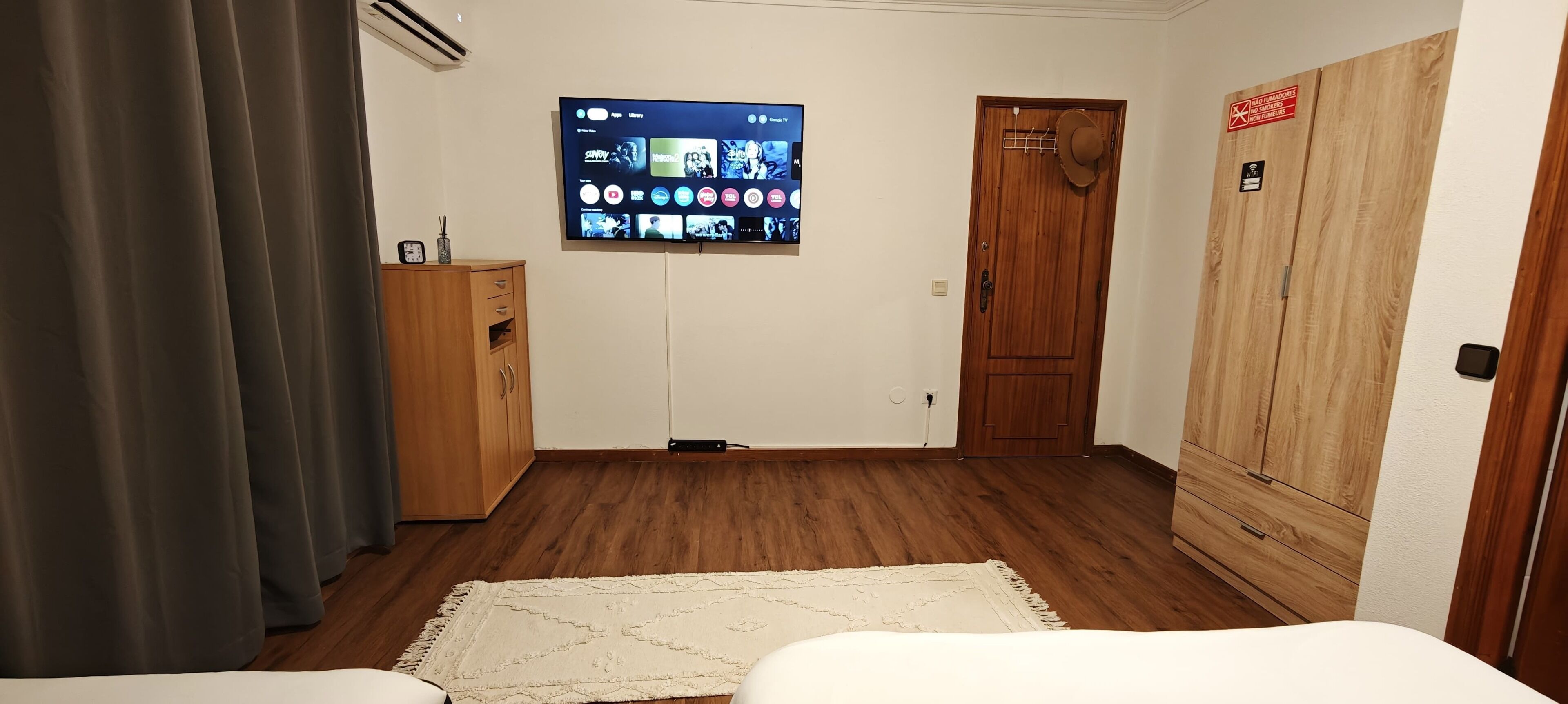 Ein Hotelzimmer mit einem Holzschrank, einem an der Wand befestigten Fernseher, der verschiedene Sender zeigt, einer kleinen Kommode und einem weißen Bett mit weißer Bettdecke.