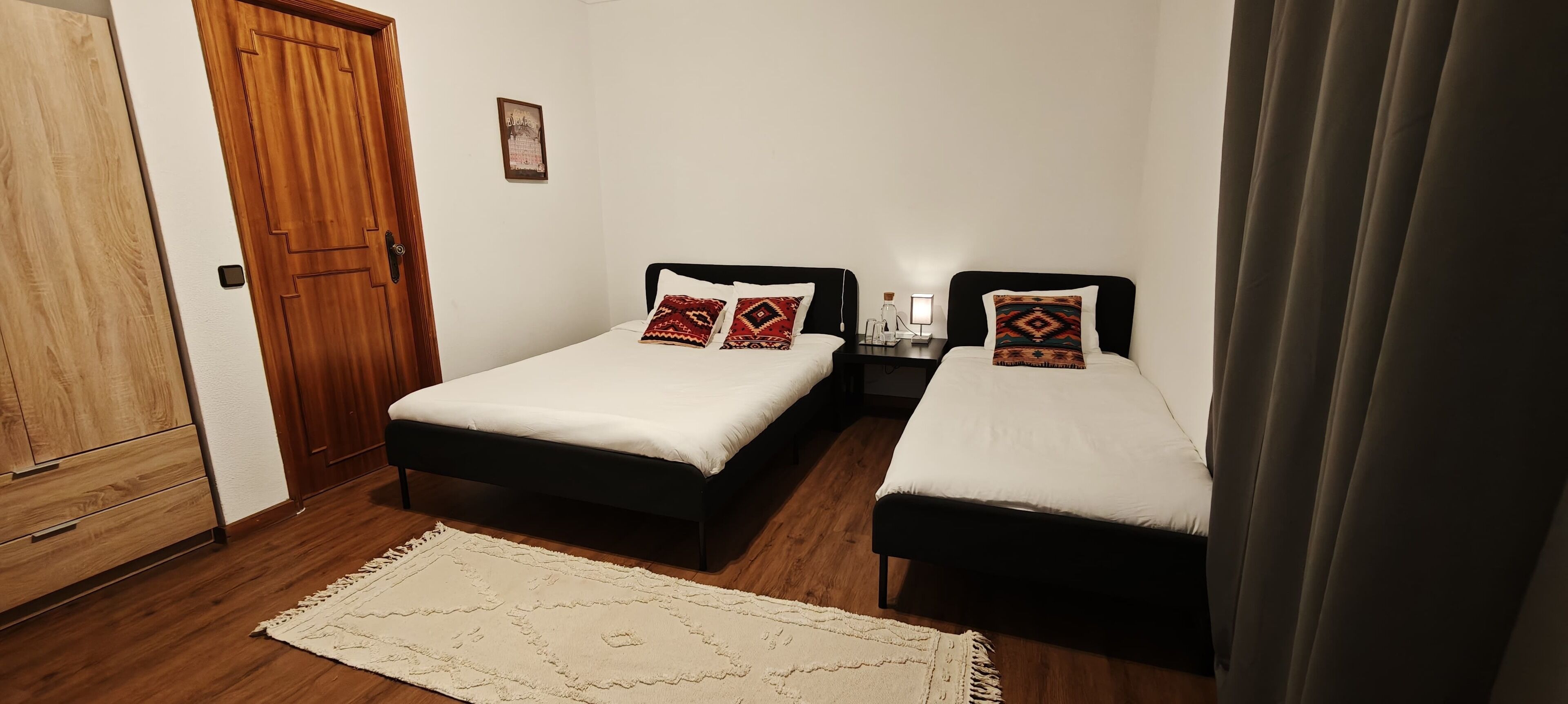 A room with two single beds, a wooden wardrobe, a small table with a lamp, and a rug on the floor.