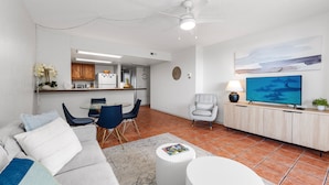Un salon moderne avec un canapé, une table basse et un téléviseur. On aperçoit une cuisine à l’arrière-plan, avec des placards et un réfrigérateur.