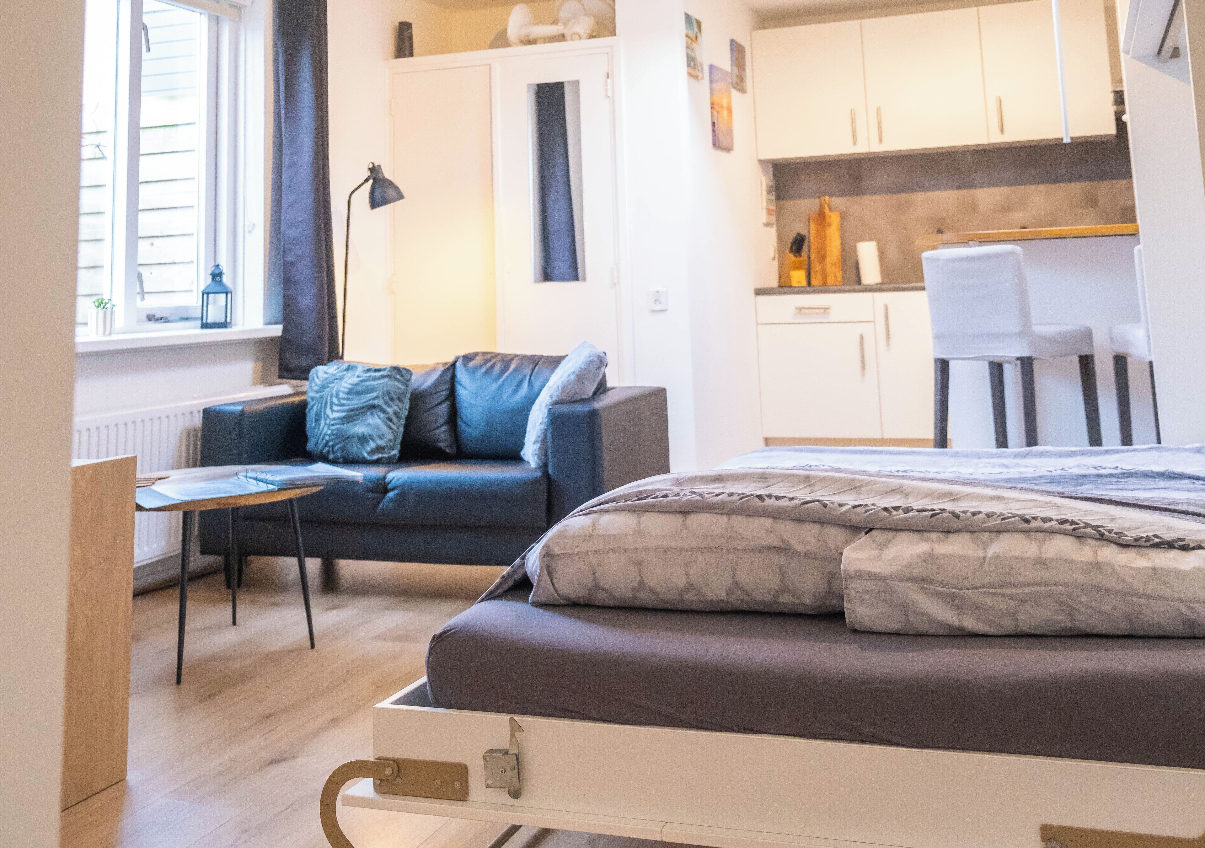 A compact bedroom with a bed, a sofa, a small table, and a kitchenette in the background.