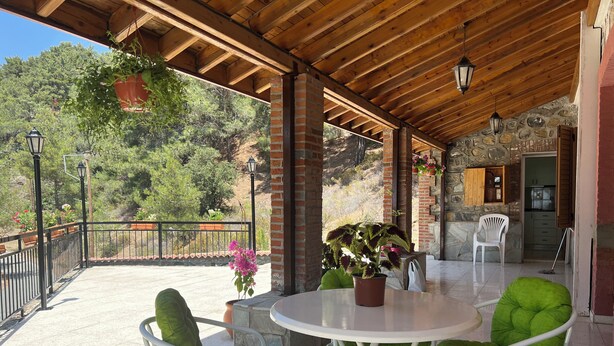 An outdoor patio area with a round table and green cushioned chairs, a brick wall, and a stone staircase leading to a kitchen area. - Charming Villa in Pano Platres, Cyprus - Your Home Away From Home (Platres)