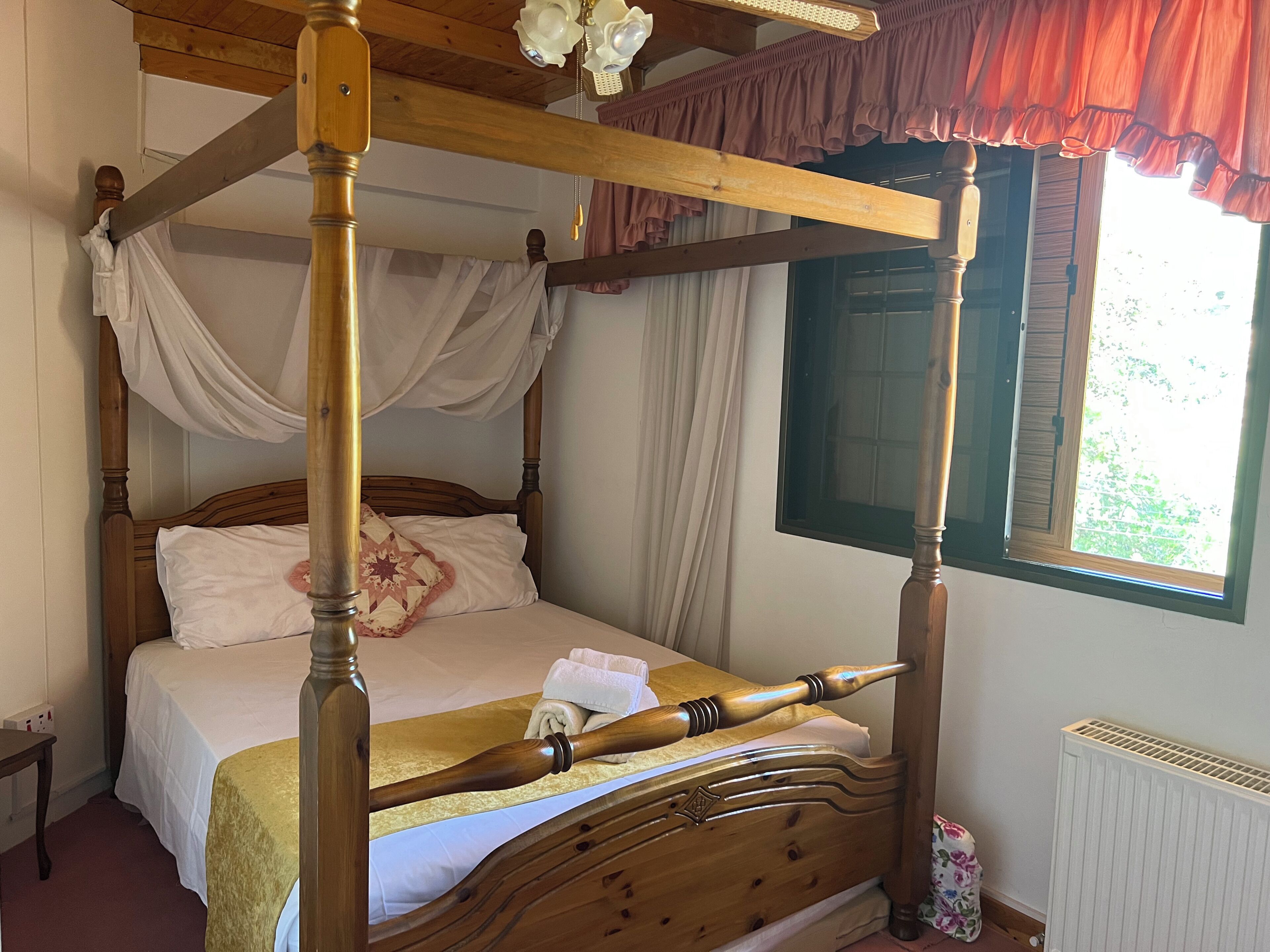 A four-poster bed with a canopy, a wooden headboard, and a window with a view of greenery.