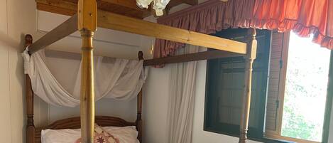 A four-poster bed with a canopy, a wooden headboard, and a window with a view of greenery.