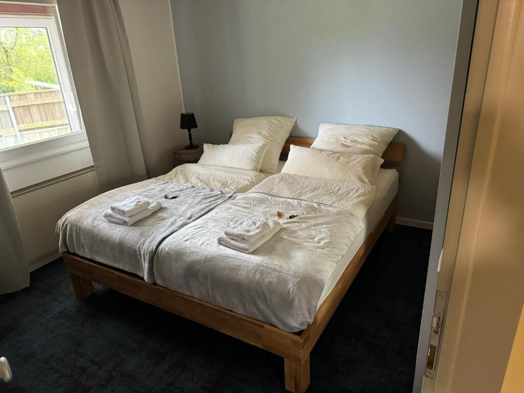 A neatly made double bed with white linens and towels, a bedside table with a lamp, and a window with curtains.