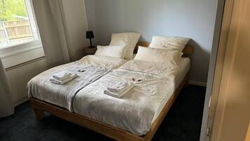 A neatly made double bed with white linens and towels, a bedside table with a lamp, and a window with curtains.