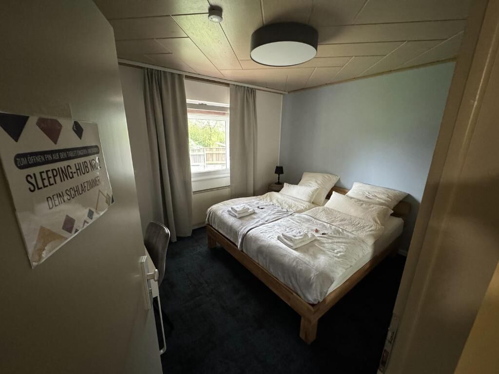 Una camera d'albergo con un letto, una finestra con tende, un cartello al muro e una lampada a soffitto.