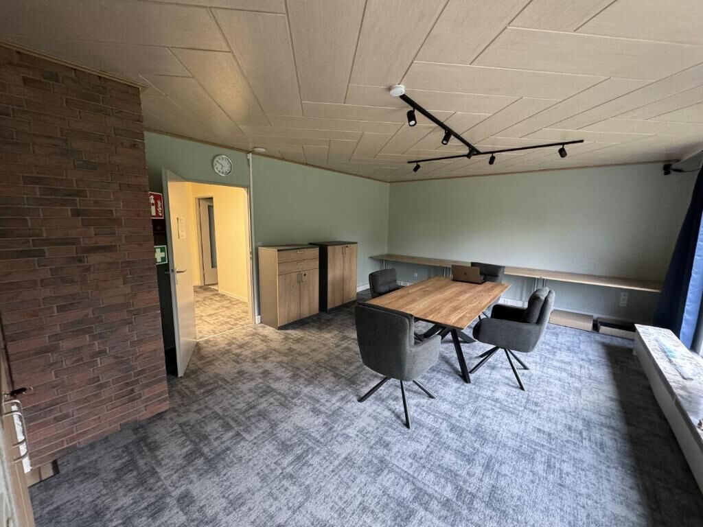 Een vergaderruimte met een houten tafel, vier stoelen, een bakstenen muur, een deur en een klok aan de muur.