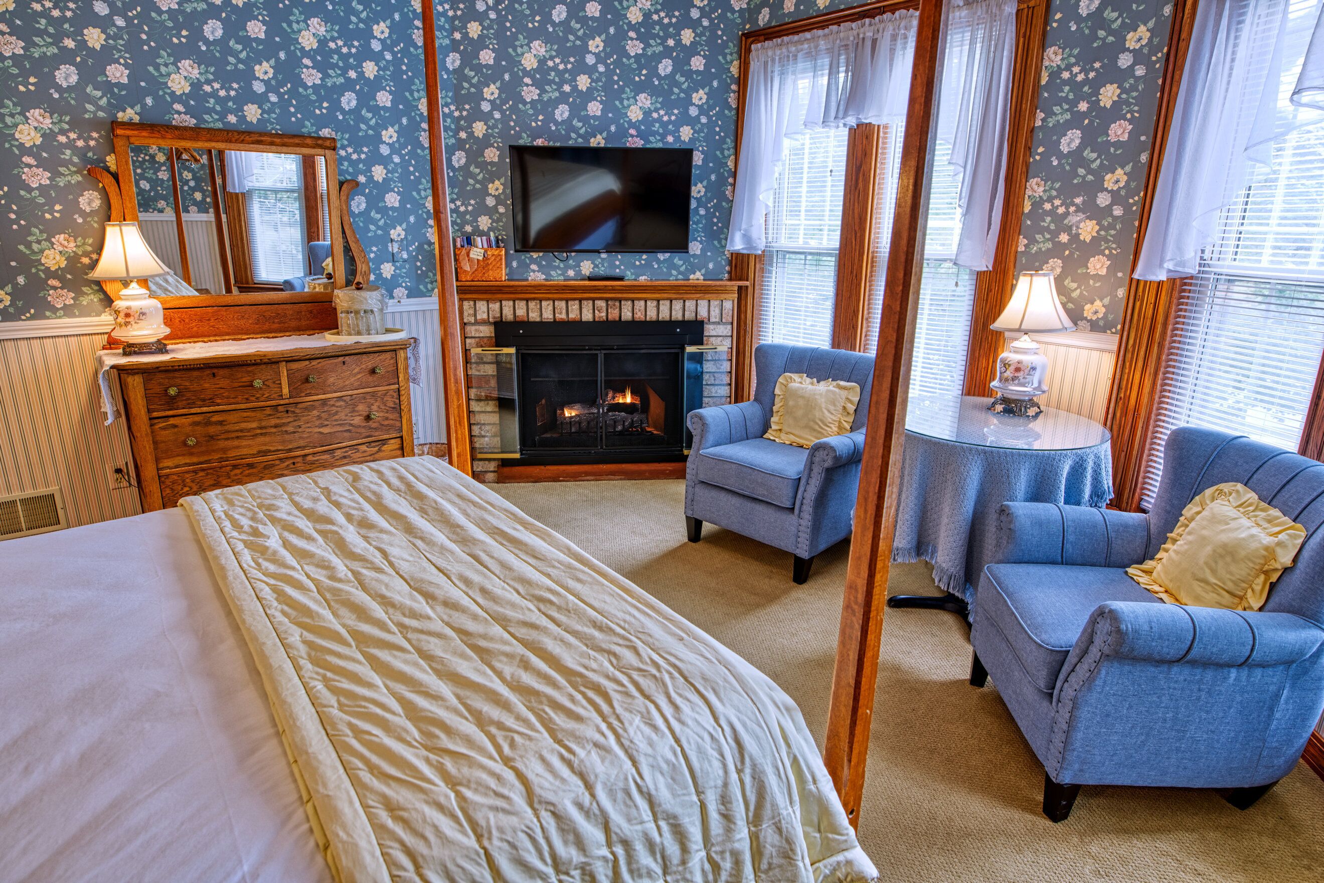 A bedroom with a bed, dresser, TV, fireplace, and two armchairs.
