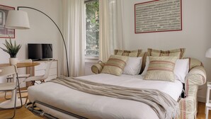 A bedroom with a bed, bedside table, lamp, TV, and a window with curtains. - JOIVY Studio Near Pinacoteca Brera (Milan)