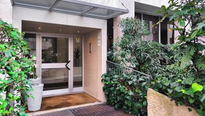 A hotel entrance with a glass door, a doormat, and a small garden area with potted plants and a stone wall. - JOIVY Studio Near Pinacoteca Brera (Milan)