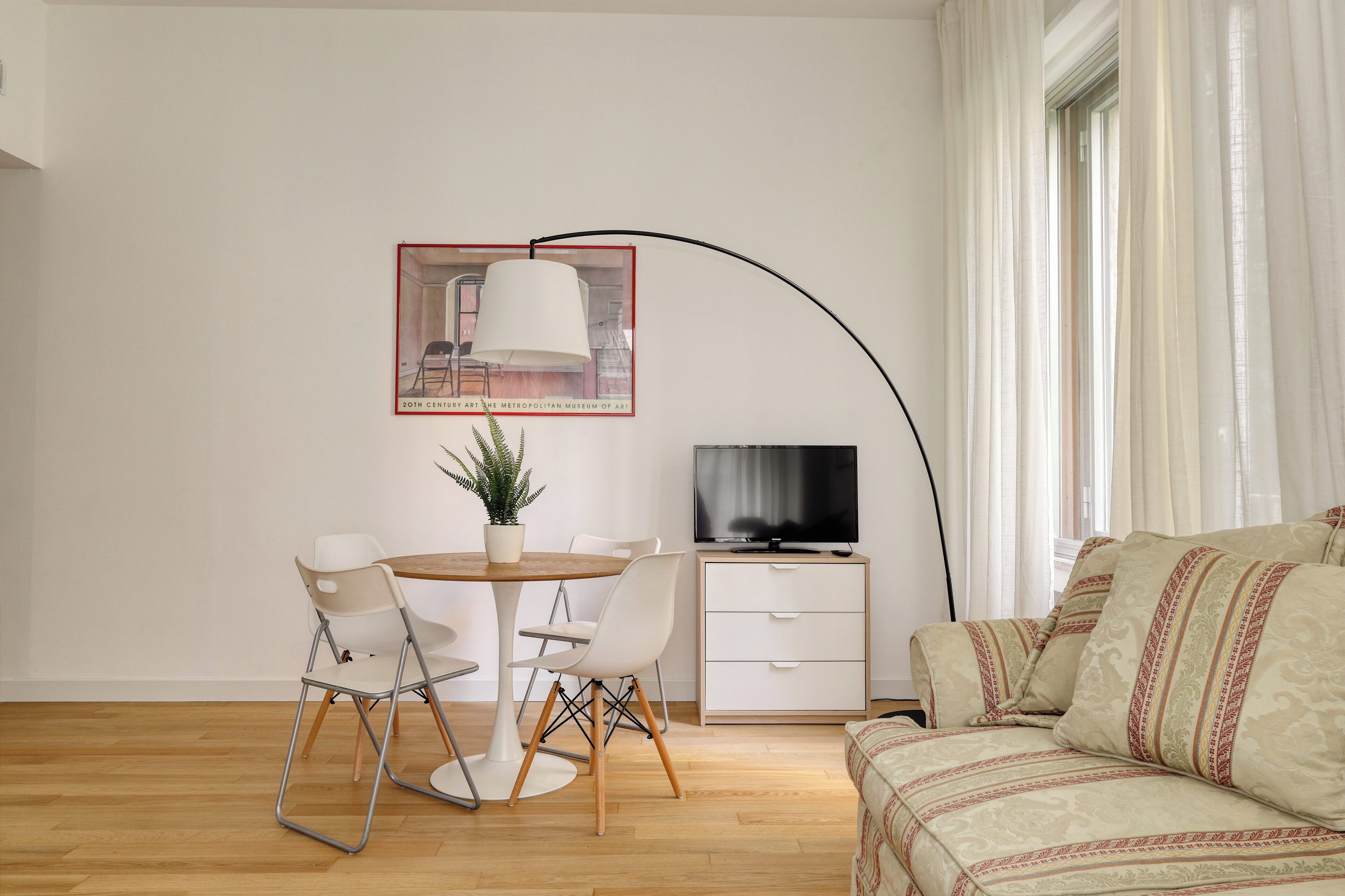 Un salón moderno con una mesa redonda de madera, sillas blancas, una planta en maceta, un televisor de pantalla plana y un sofá con cojines estampados.