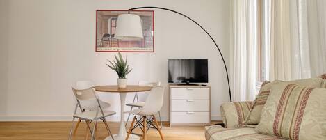 Un salón moderno con una mesa redonda de madera, sillas blancas, una planta en maceta, un televisor de pantalla plana y un sofá con cojines estampados.