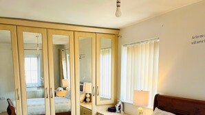 A bedroom with a wooden bed, a wardrobe with a mirror, a bedside table with a lamp, and a window with blinds.