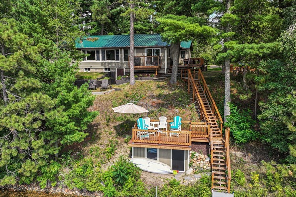 A charming lakeside retreat nestled among towering pines, featuring multi-level decks and wooden stairs connecting the main cabin to waterfront access.