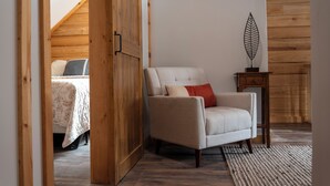Une pièce avec un plafond en bois, un lit recouvert d’une couvre-lit à motifs floraux, un fauteuil beige et une petite table d’appoint en bois ornée de motifs végétaux.