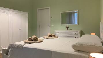 Een net opgemaakt bed met opgerolde handdoeken, een bedlampje en een dressoir met een spiegel erboven, in een kamer met groene wanden en witte meubels.
