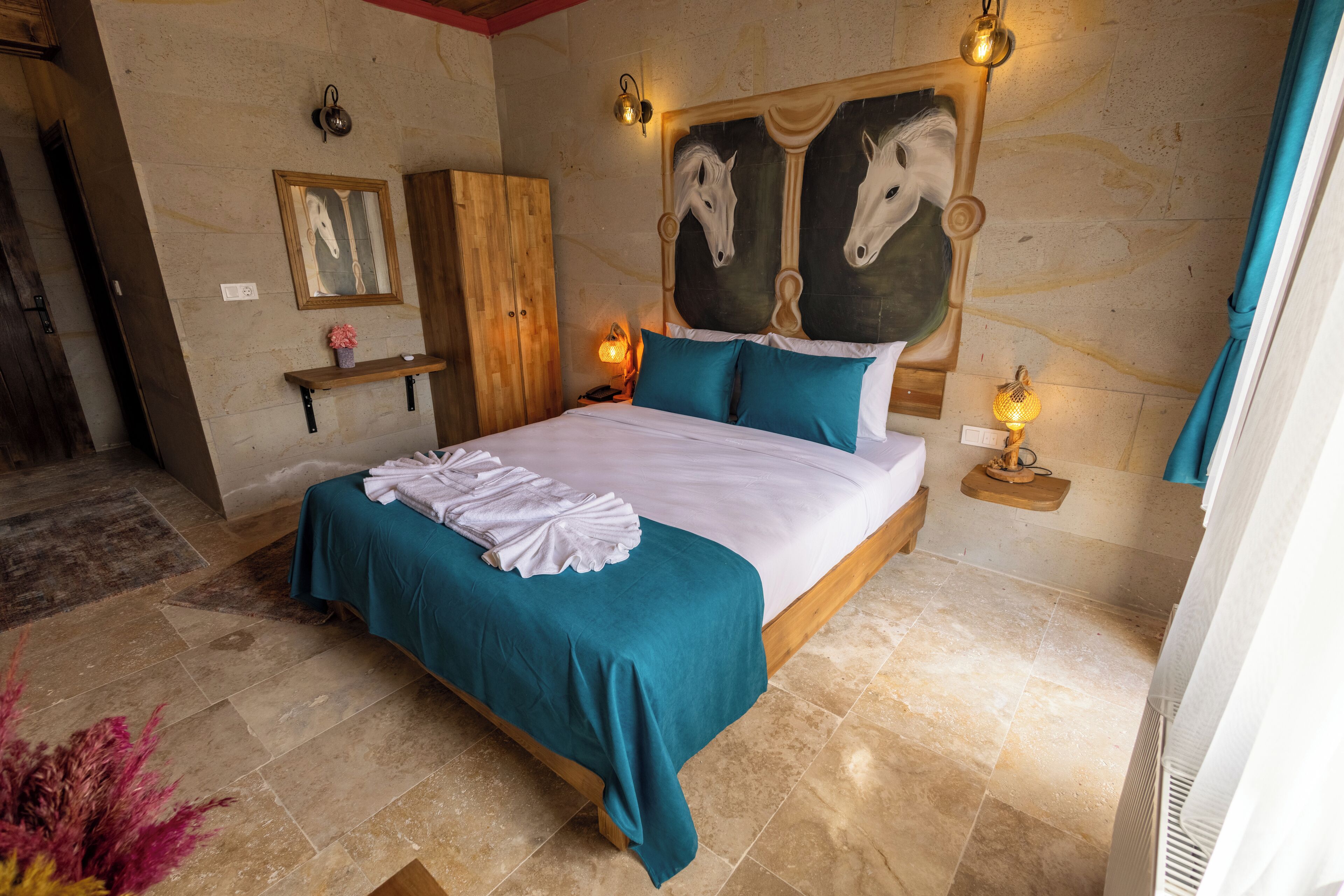 A bedroom with a large bed, teal bedding, a wooden headboard with horse head decorations, a nightstand with a lamp, and a small shelf with a framed picture.