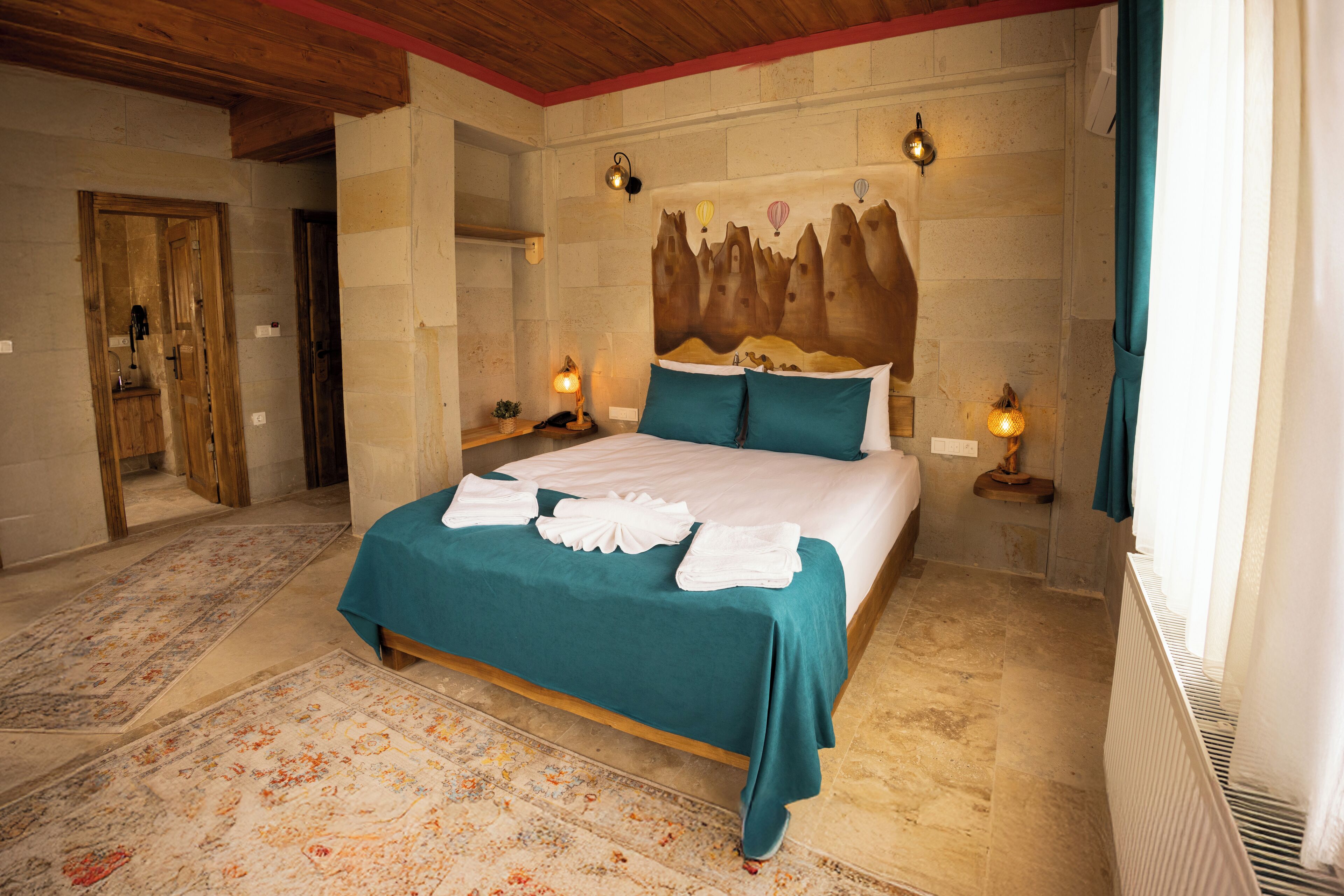 A bedroom with a bed, two bedside lamps, a headboard with a mountain design, and a large window with curtains.