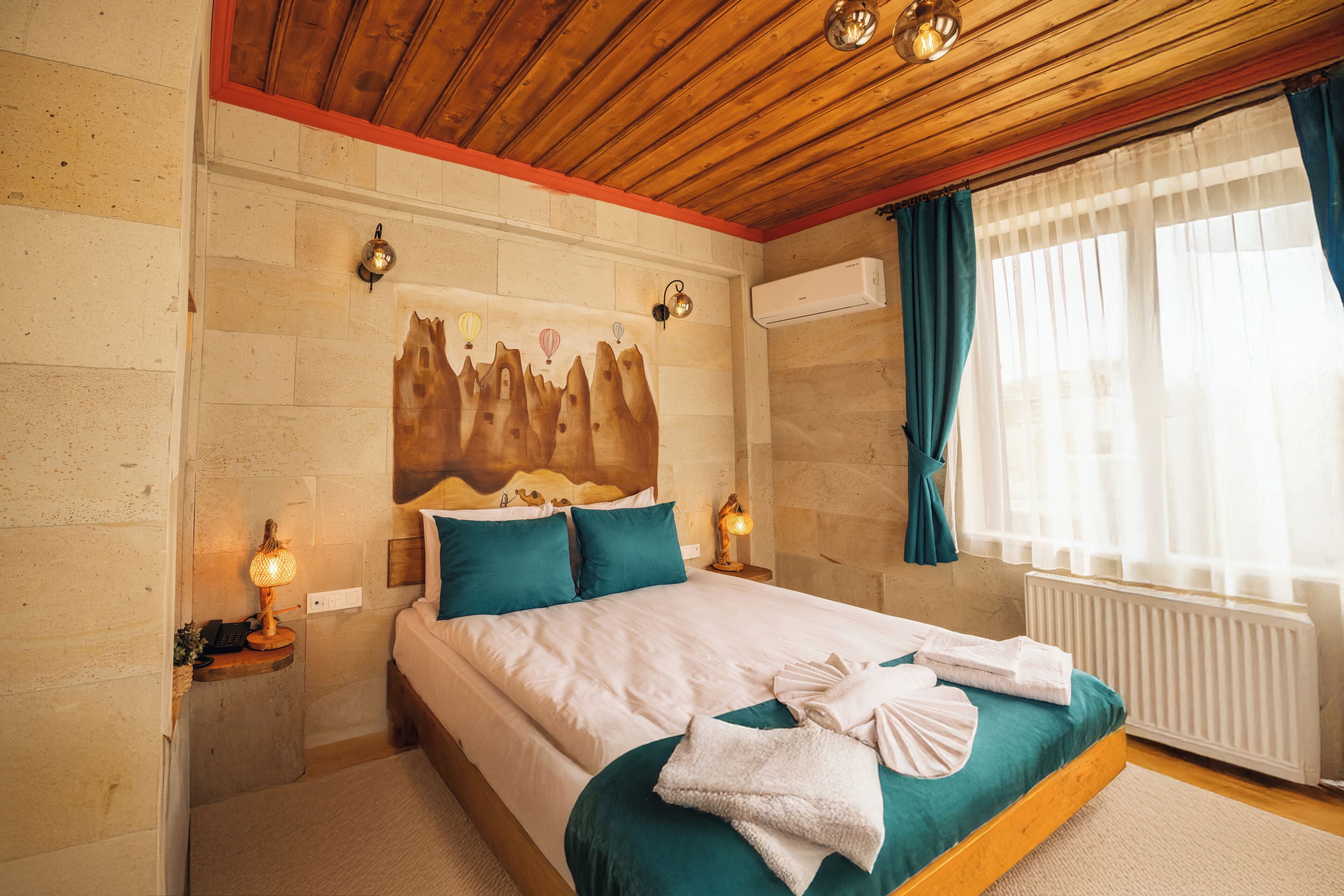 A hotel room with a wooden ceiling, a stone wall, a bed with teal bedding, two bedside lamps, and a window with curtains.