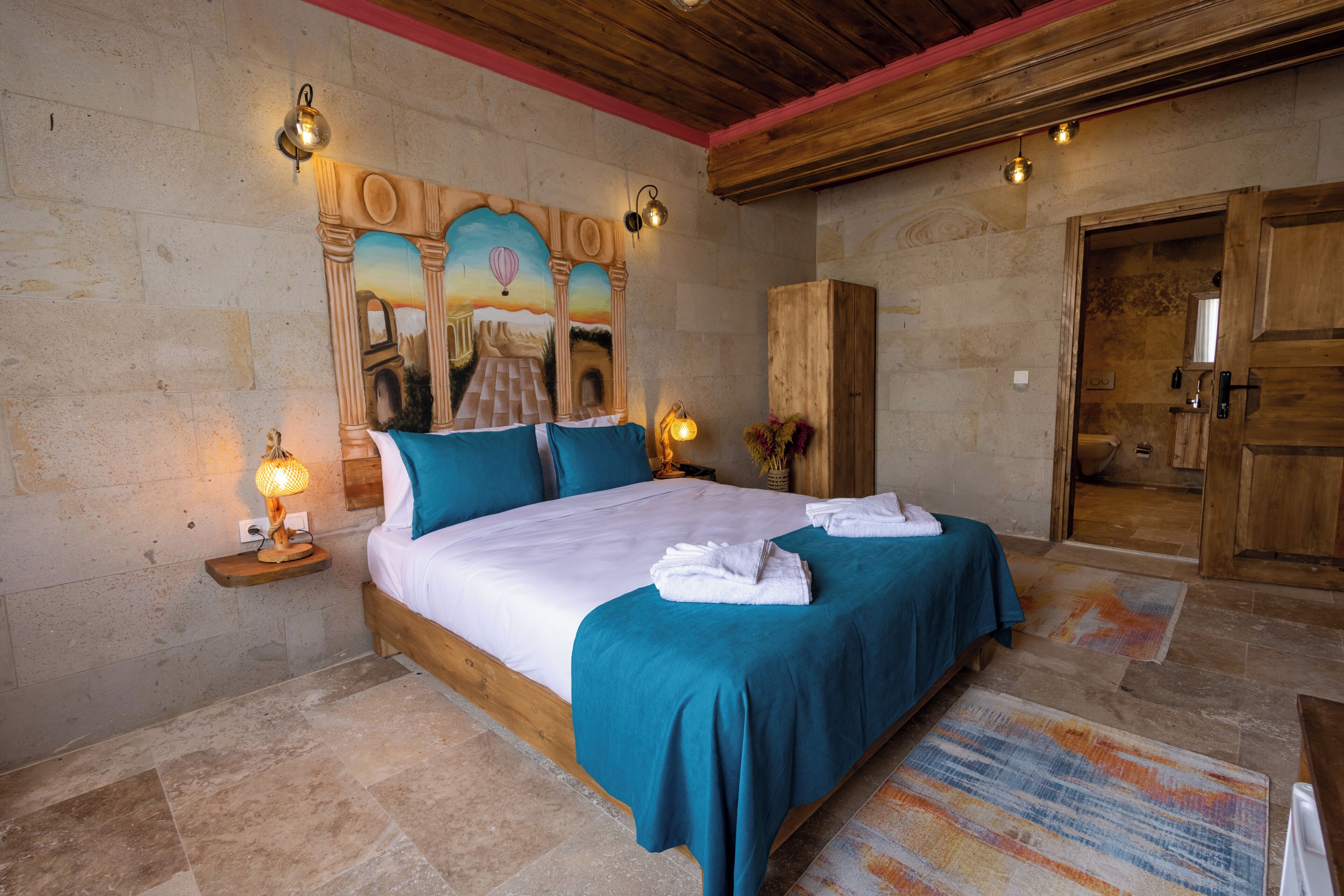 A bedroom with a large bed, wooden headboard, and a mural on the wall. There are two bedside tables with lamps and a wooden wardrobe.