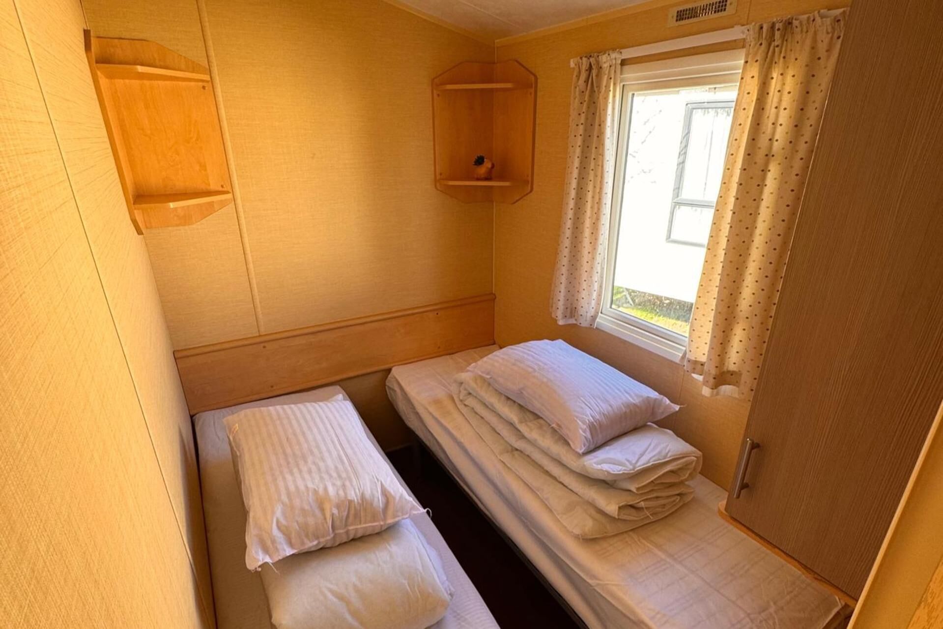 A small, single-bed room with a wooden headboard, a window with curtains, and a shelf above the bed.