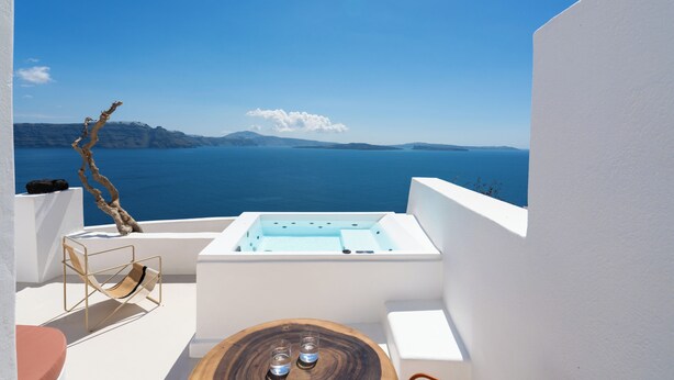 Un balcón con una bañera blanca, una mesa de madera con vasos, una silla y vistas al mar y a las montañas.