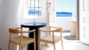 Una habitación con una mesa redonda, dos sillas de mimbre, un ventanal con vistas al mar y una puerta que da al exterior.