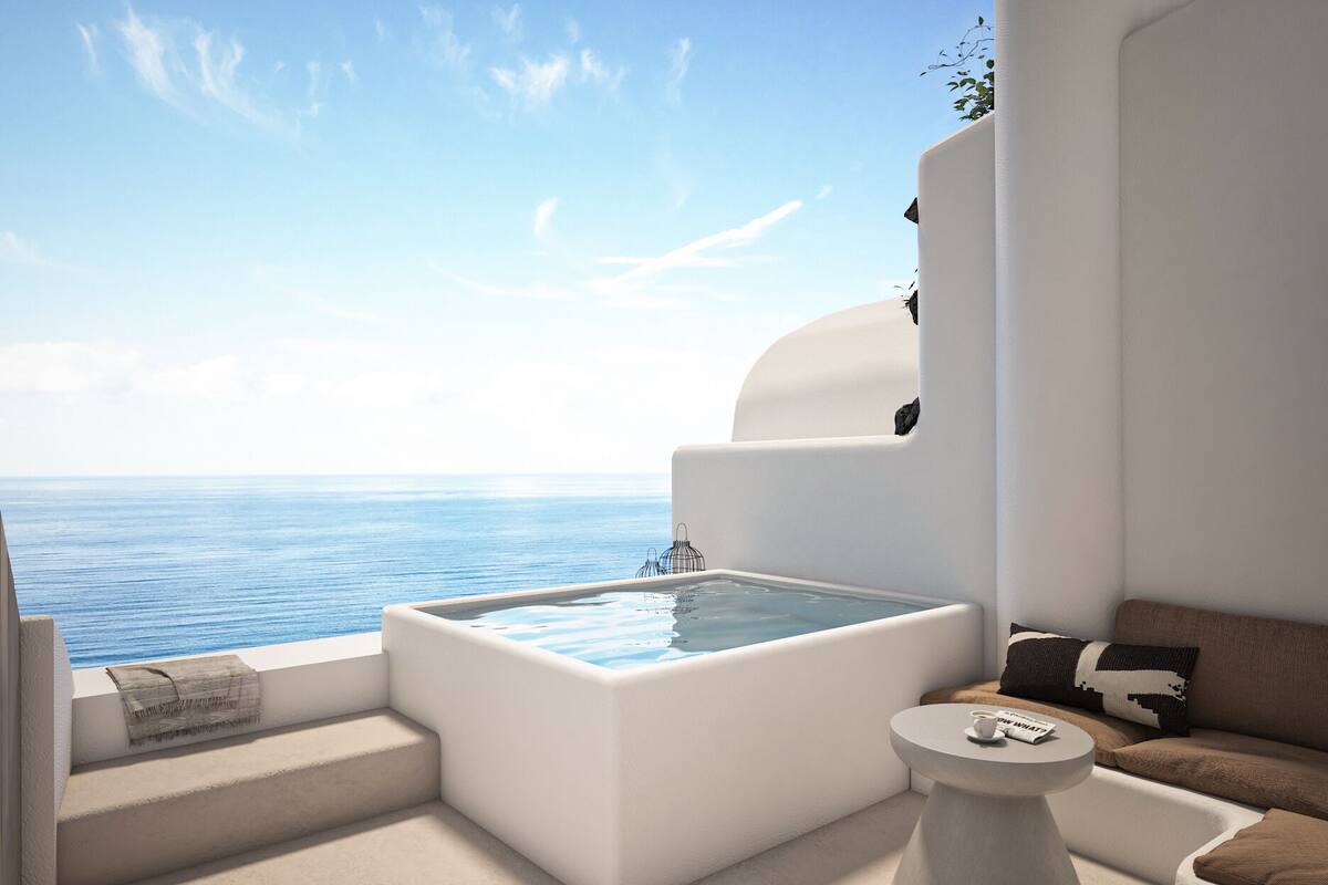 A modern, minimalist room with a large, square hot tub overlooking the sea, a small round table, and a sofa with cushions.