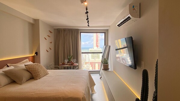 A hotel room with a bed, a small dining table with a plant, a TV mounted on the wall, and a window with a city view. - Simple by Hosppedar (João Pessoa)