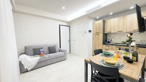 Ein kompakter Wohnbereich mit Küchenzeile, Essplatz und grauem Sofa mit Zierkissen.