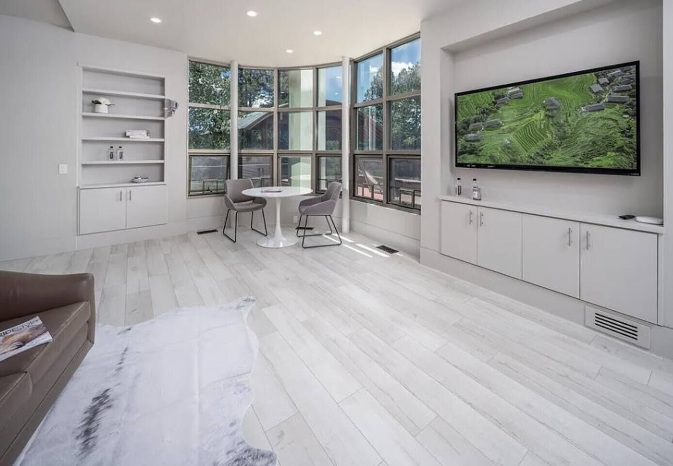 A modern living room with a large TV, white cabinetry, a round dining table, and a large window offering a view of the outdoors.