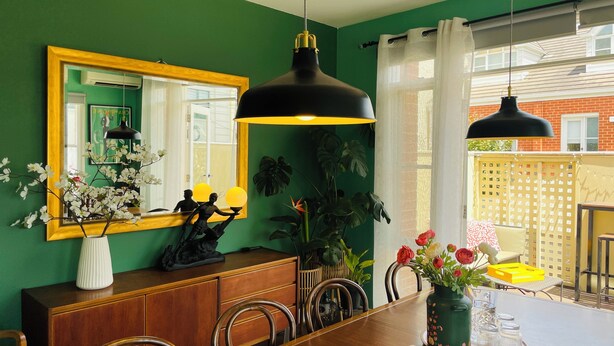 Comedor con mesa de madera, sillas, espejo con marco dorado, lámpara colgante negra y un jarrón con flores.