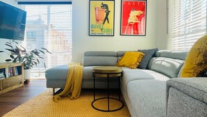 Un salón con un sofá gris, cojines amarillos, una alfombra estampada y dos carteles en la pared.