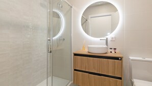 A modern bathroom with a glass-enclosed shower, a round mirror with a light, and a wooden vanity with a sink.