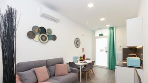 A modern living room with a grey sofa, a dining table with chairs, a kitchen area with a toaster, and decorative wall art.