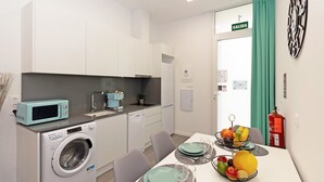 Une cuisine compacte équipée d’un lave-linge, d’un four à micro-ondes et d’un évier. On y trouve également un coin repas avec une table dressée pour deux personnes, et un panier de fruits posé sur la table.