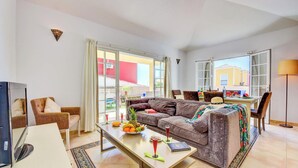 A spacious living area with a large sofa, a dining table with a fruit bowl and drinks, and a TV on a stand.