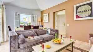 A living room with a grey sofa, a dining table with fruit and drinks, and a view of the outside.
