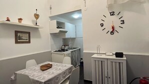 Kompakti keittiö, jossa on valkoiset kaapit, ruokapöytä tuoleineen, seinäkello ja pieni kaappi, jonka sisällä on palosammutin.