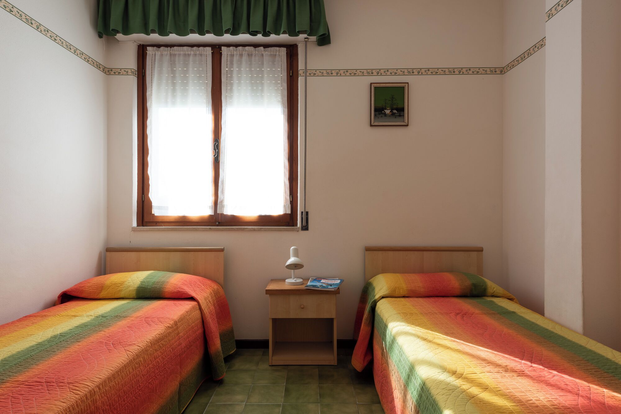 A small, well-lit bedroom with two single beds, a nightstand, a window with curtains, and a framed picture on the wall.