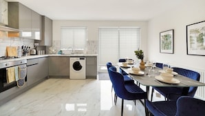 Une cuisine moderne équipée d’un lave-linge intégré, un coin repas avec des chaises bleues et une grande fenêtre avec des stores.