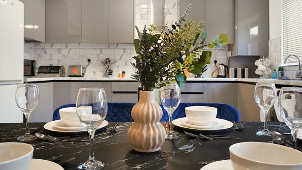 Une cuisine moderne avec des meubles gris, un plan de travail en marbre et un coin repas agrémenté d’assiettes, de bols et de verres à vin blancs.
