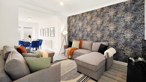 Un salon moderne avec un canapé d’angle, un coin repas avec des chaises bleues et un grand mur décoré d’une œuvre abstraite.