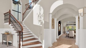 A grand staircase with a wooden finish and black balusters, leading to a spacious hallway with white columns and a view of an outdoor patio.