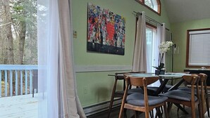 A dining area with a wooden table and chairs, large windows with curtains, and a colorful abstract painting on the wall.