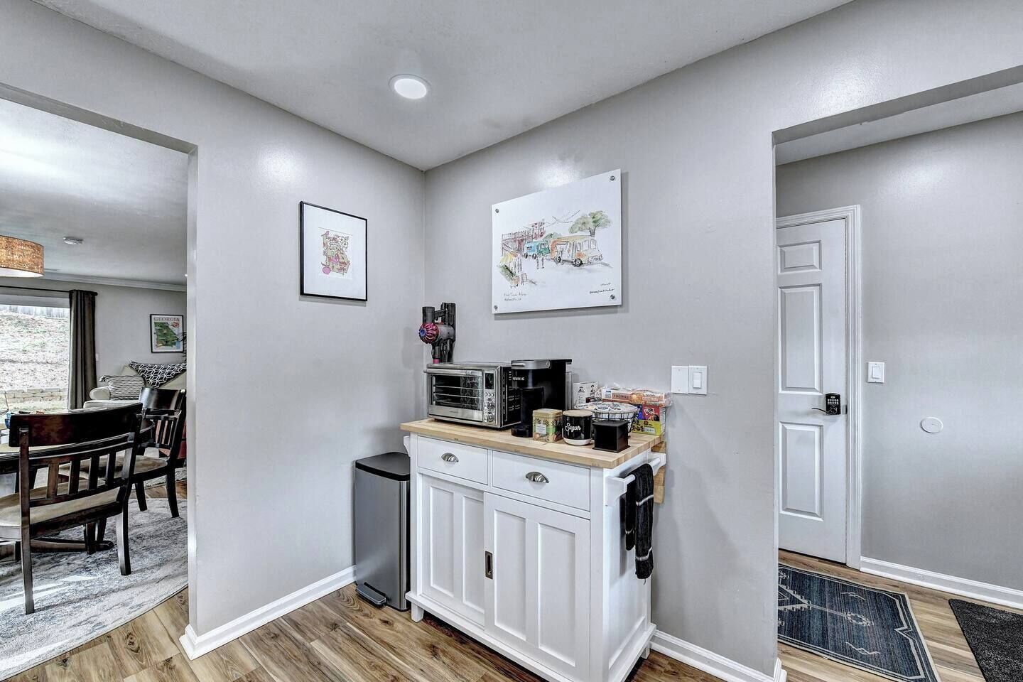 A compact kitchen with white cabinetry, a microwave, a toaster, and a coffee maker. There is a small trash bin and a door with a keypad lock.