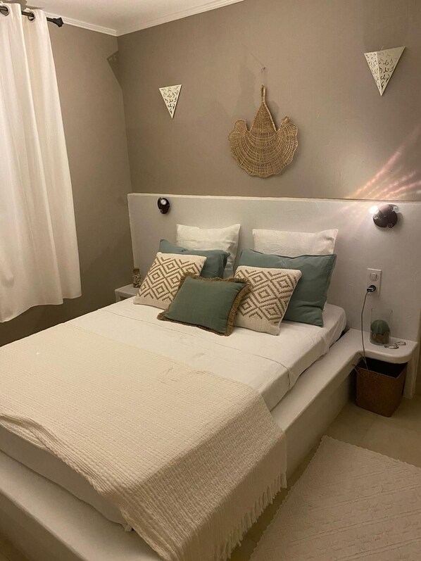 A neatly made bed with a white and beige bedspread, two pillows with geometric patterns, and a decorative woven piece on the wall.